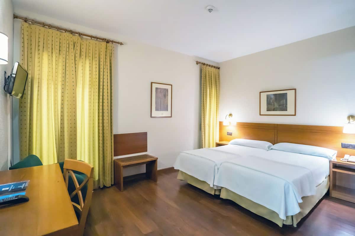A hotel room with a wooden bed, a desk with a chair, a bench, and two framed pictures on the walls.