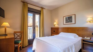 Una habitación de hotel con cama, escritorio, silla, ventana con cortinas, lámpara de pared y un cuadro enmarcado.