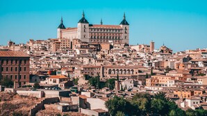 Un paisaje urbano histórico con un edificio imponente que recuerda a un castillo, con torres, rodeado de construcciones densamente agrupadas y diversos estilos arquitectónicos.