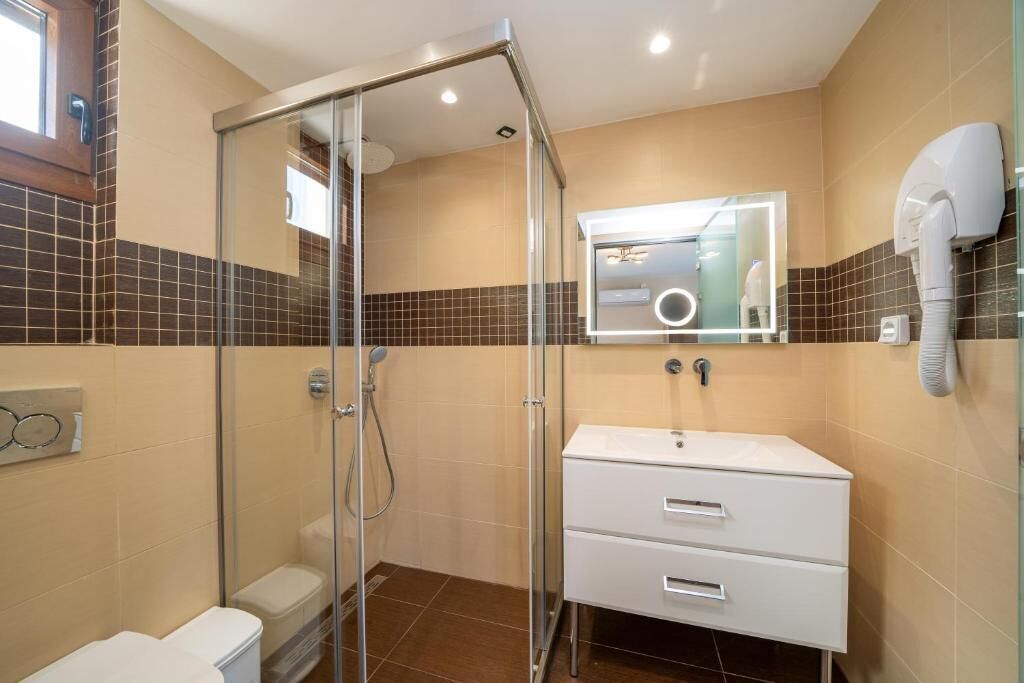 Un bany modern amb dutxa tancada amb vidre, un lavabo blanc i un mirall amb llum integrada.