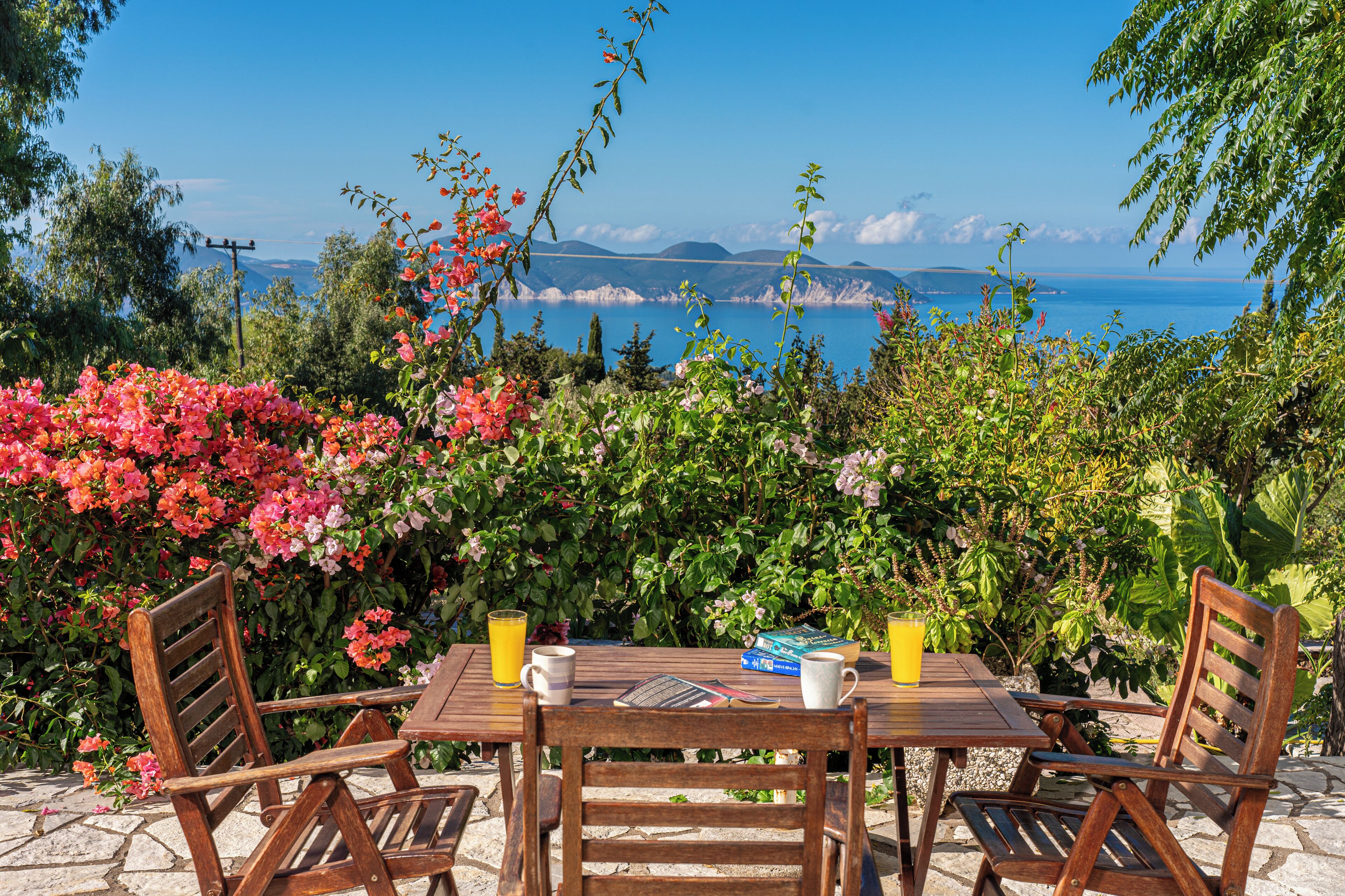 Una mesa de madera con sillas, un libro y una taza, frente a un fondo de flores en flor y vista al mar.