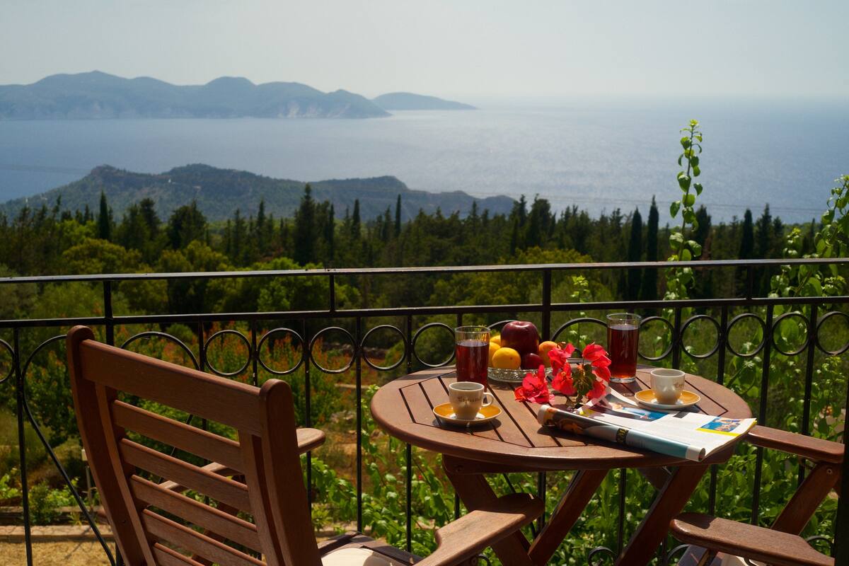 Una mesa de madera con un vaso de jugo, una taza de café y un libro en un balcón con vistas a un paisaje montañoso.