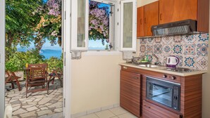 A compact kitchen with wooden cabinets, a built-in oven, and a window overlooking a garden with purple flowers. - Panoramic Sunset (Kefalonia)