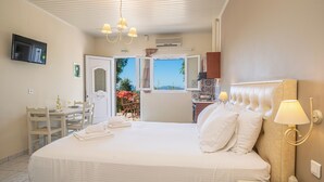 A bedroom with a large bed, a TV mounted on the wall, a dining area with chairs and a table, and a view through an open door. - Panoramic Sunset (Kefalonia)