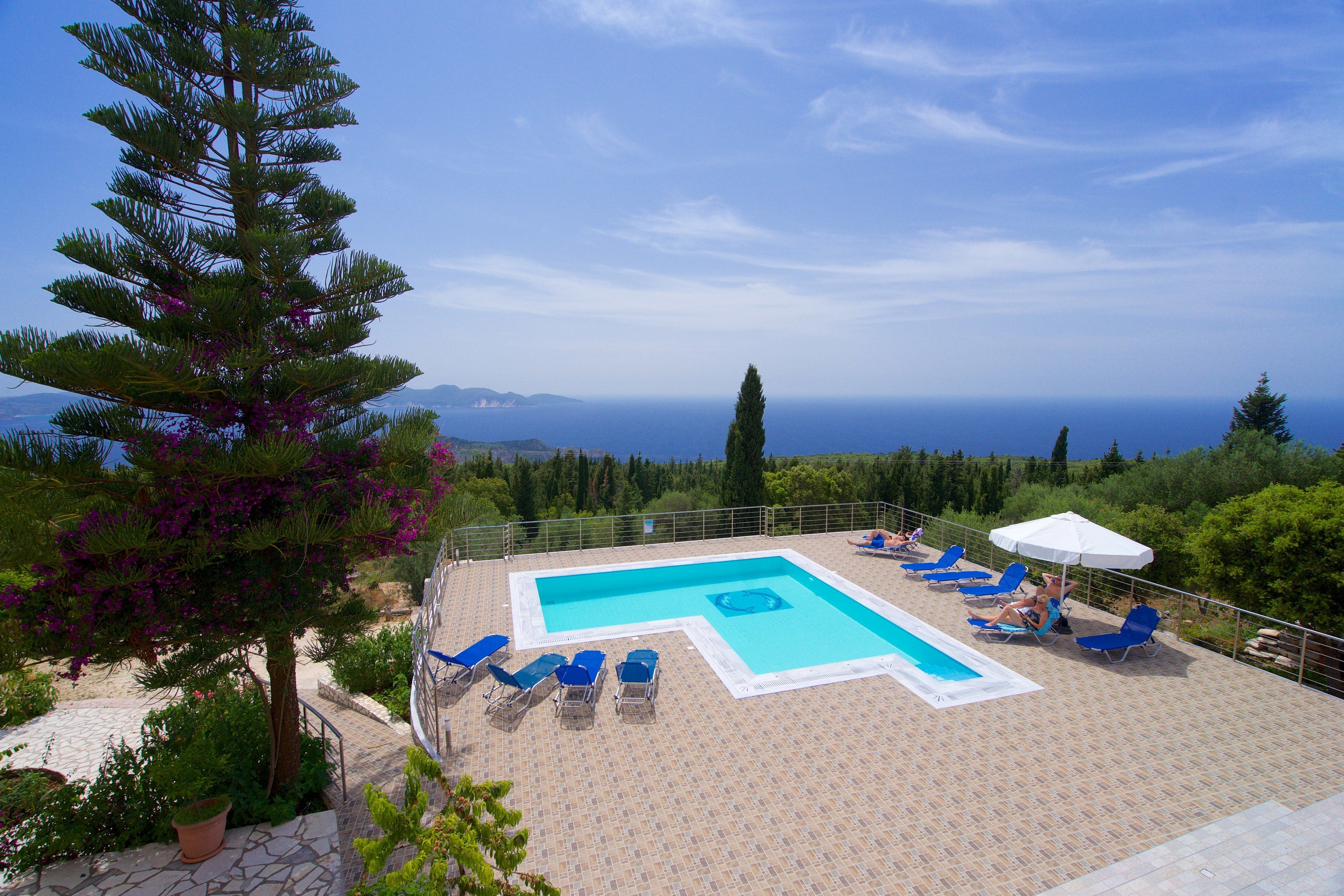 A rooftop pool with blue chairs and a white umbrella, surrounded by greenery and overlooking a scenic view.