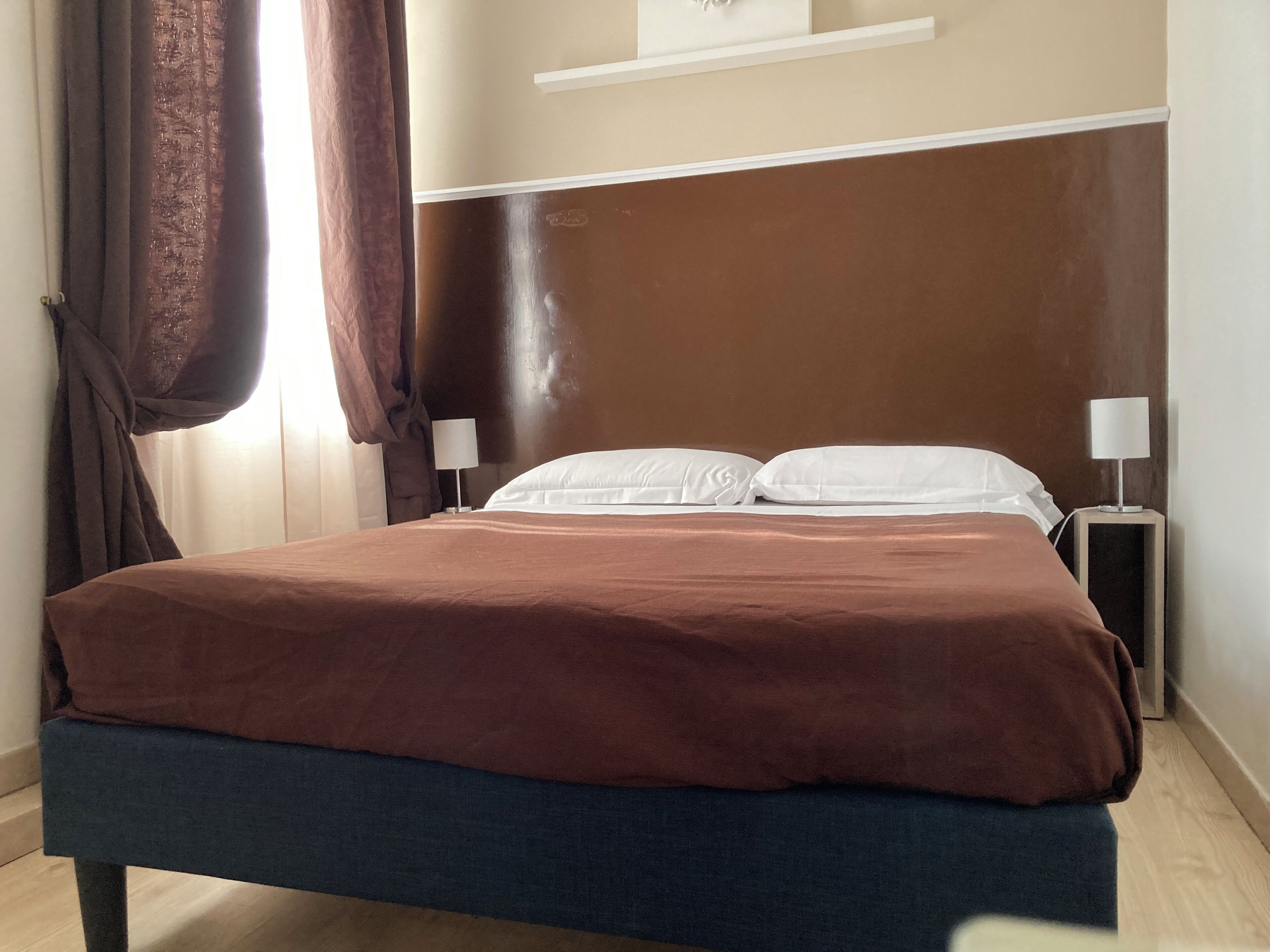 A neatly made bed with a brown headboard, two bedside tables with lamps, and a window with curtains.