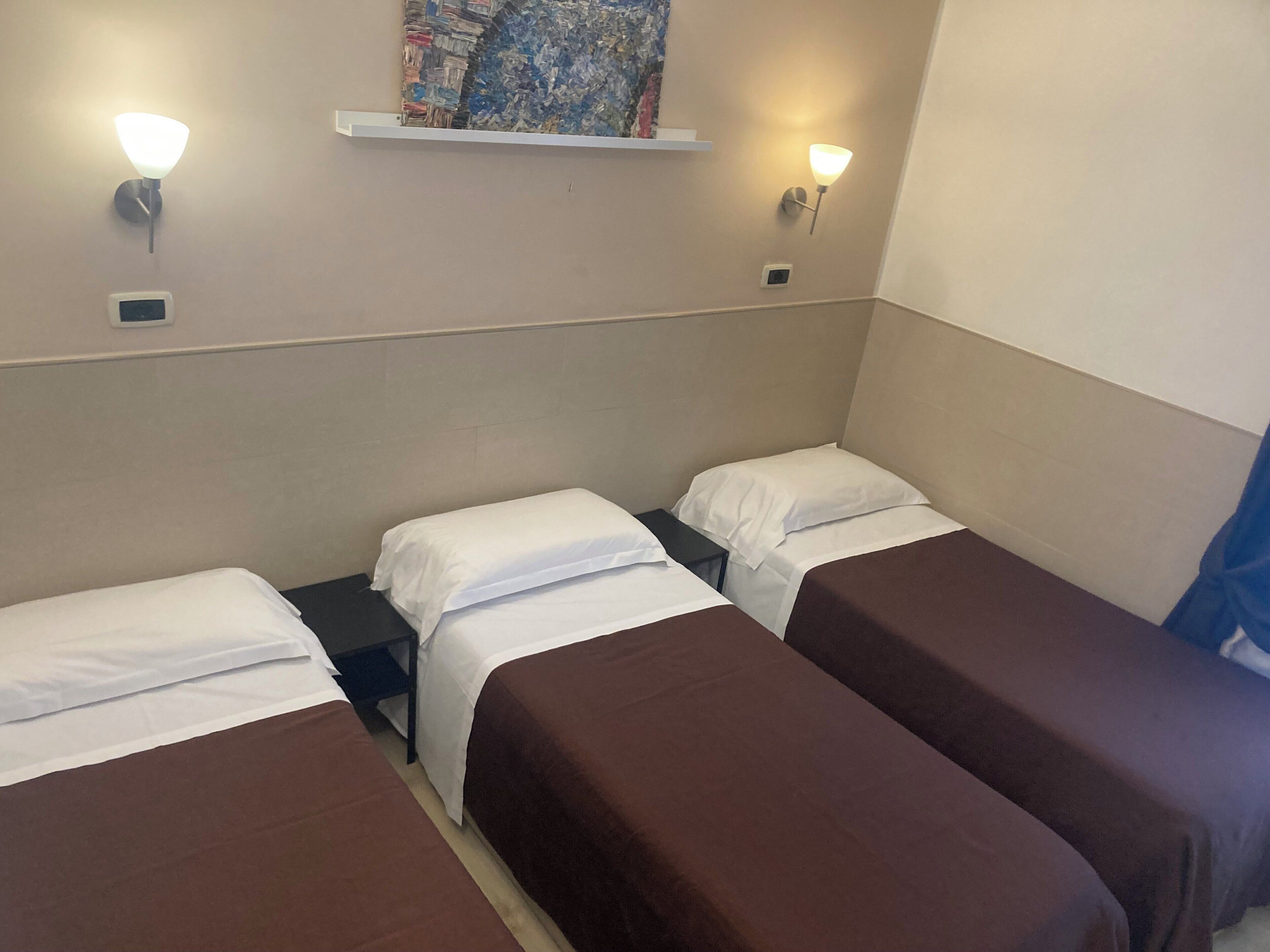 A hotel room with three single beds, each with a brown bedspread and white pillows. There are two wall-mounted light fixtures on either side of a painting.