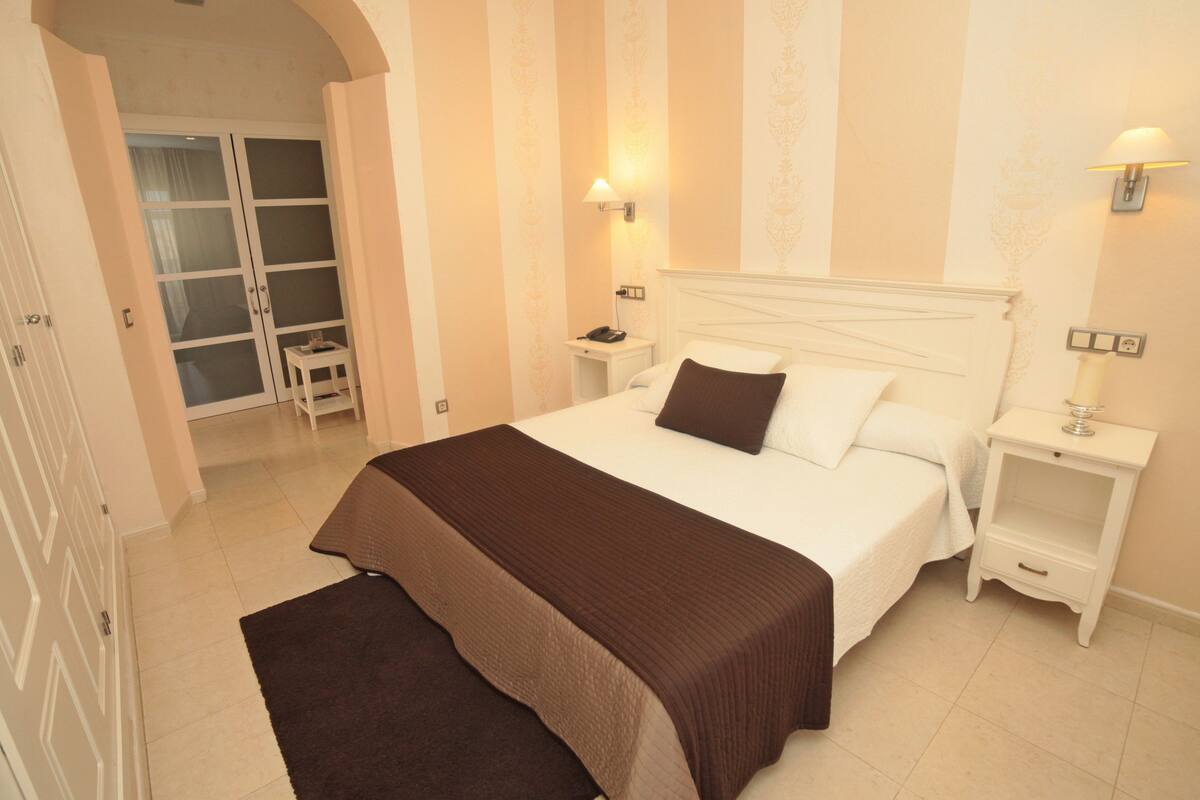 Una camera da letto con un letto grande, due comodini, un tavolino e una lampada, con una porta che dà su un bagno.