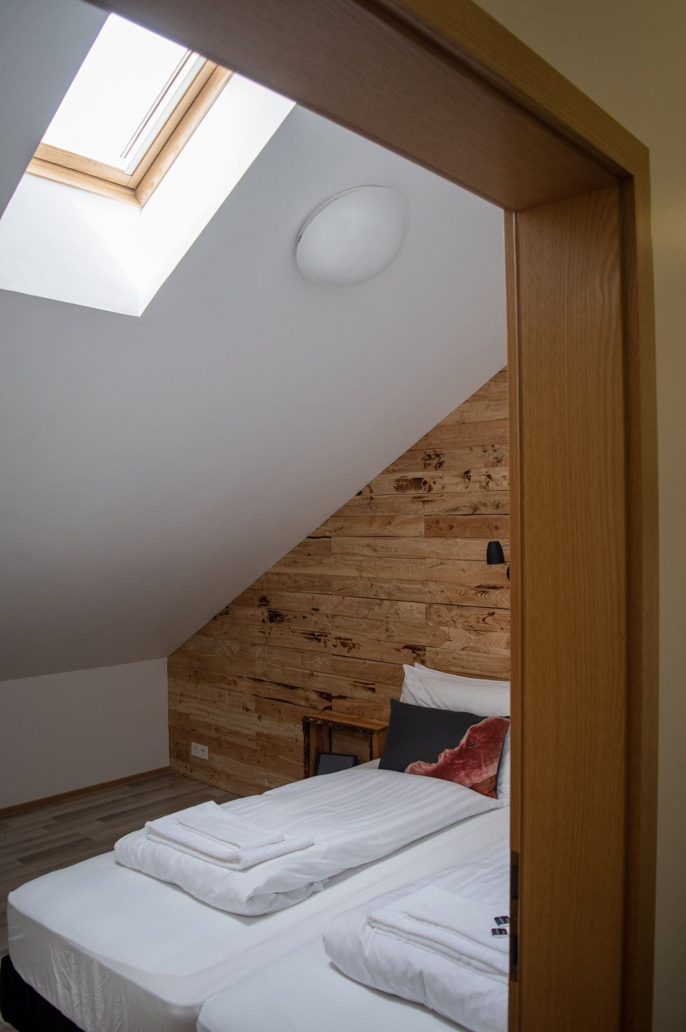 A bedroom with a sloped ceiling, wooden panel walls, a skylight, and a neatly made bed.