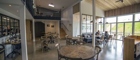 A spacious interior with a central round table surrounded by chairs, a staircase leading to an upper level, and large windows offering views of the outdoors.