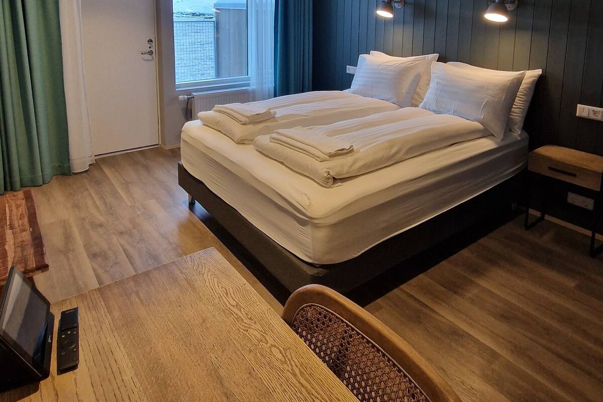 Habitación de hotel con una cama grande, un escritorio de madera, una silla, una ventana con cortinas y vistas a montañas.