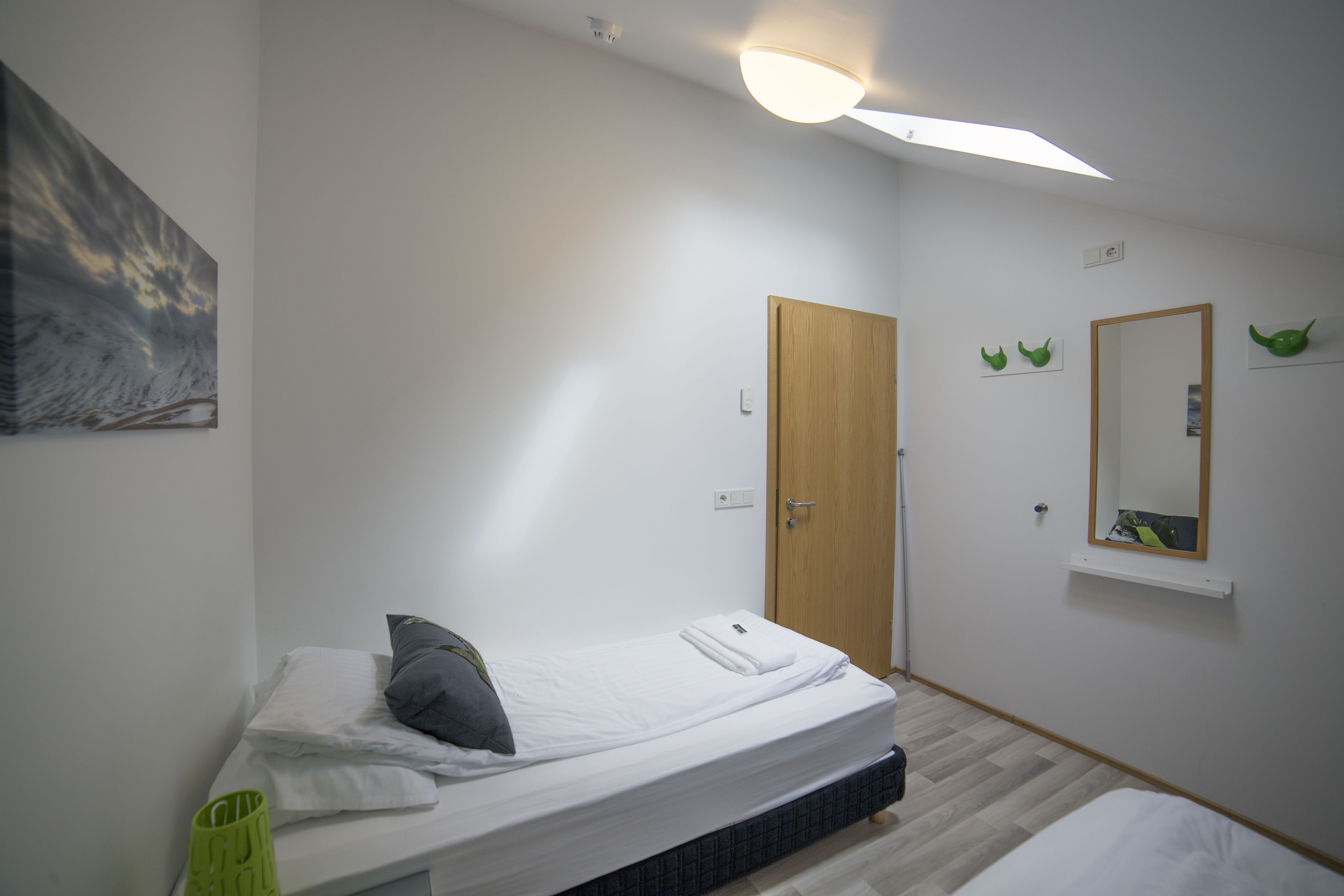 A small, clean room with a single bed, a wooden door, a mirror, and a wall-mounted shelf.