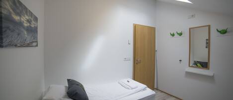 A small, clean room with a single bed, a wooden door, a mirror, and a wall-mounted shelf.