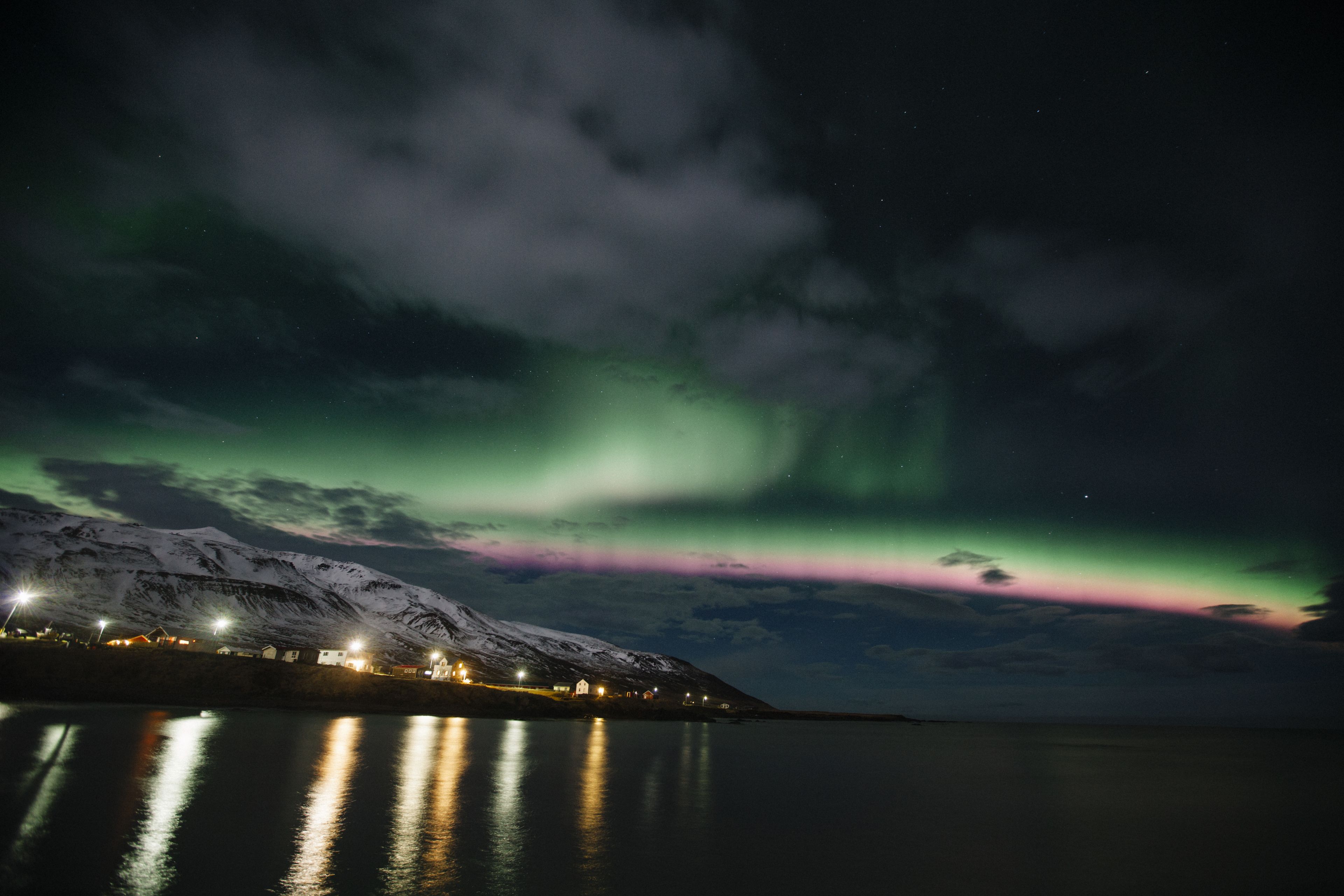 Ein verschneiter Berg mit einer Stadt entlang eines ruhigen Gewässers, das Lichter und Polarlicht am Himmel reflektiert.
