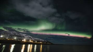 Una montaña nevada con un pueblo a lo largo de un cuerpo de agua tranquilo, que refleja luces y la aurora boreal en el cielo.