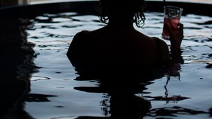 Una persona en un jacuzzi con una bebida, mirando un cuerpo de agua y montañas a través de una ventana.