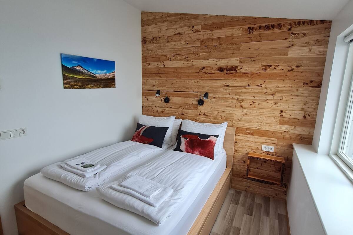 Un dormitorio con cabecera de madera, cama con ropa blanca, dos lámparas de noche y una ventana con vistas al exterior.