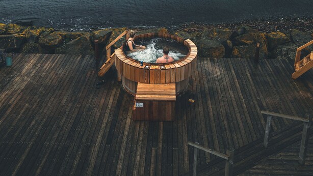 Una terraza de madera con un jacuzzi y sillas que dan a un mar tranquilo y un muelle distante.