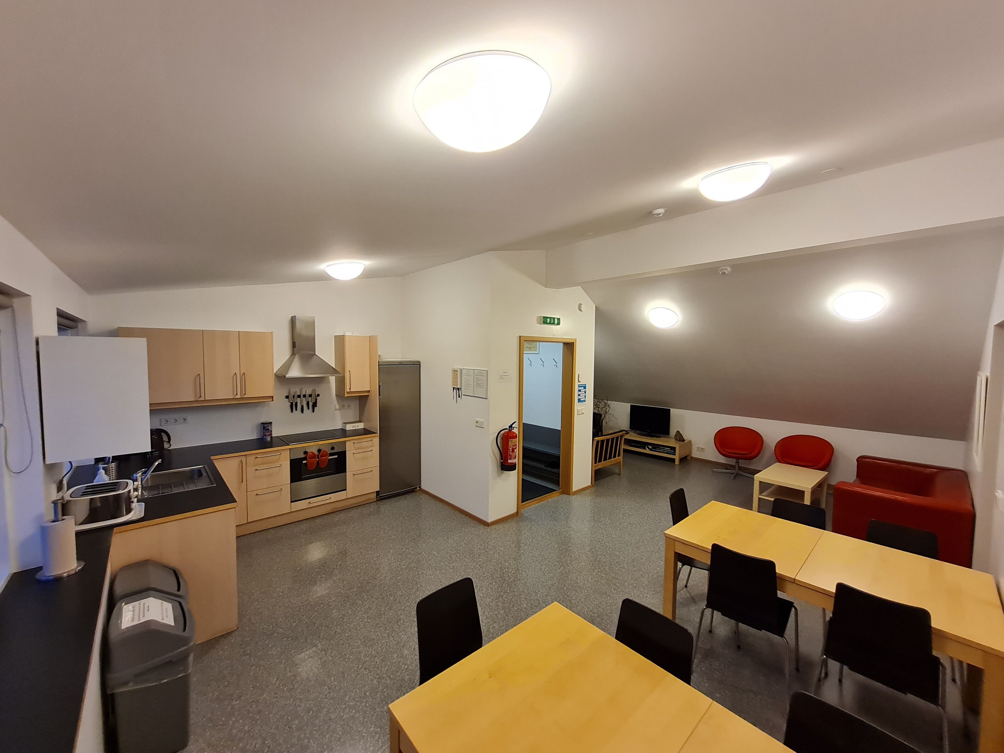 A modern kitchen with wooden cabinets, a stainless steel refrigerator, and a built-in oven. There is a dining area with yellow tables and black chairs.