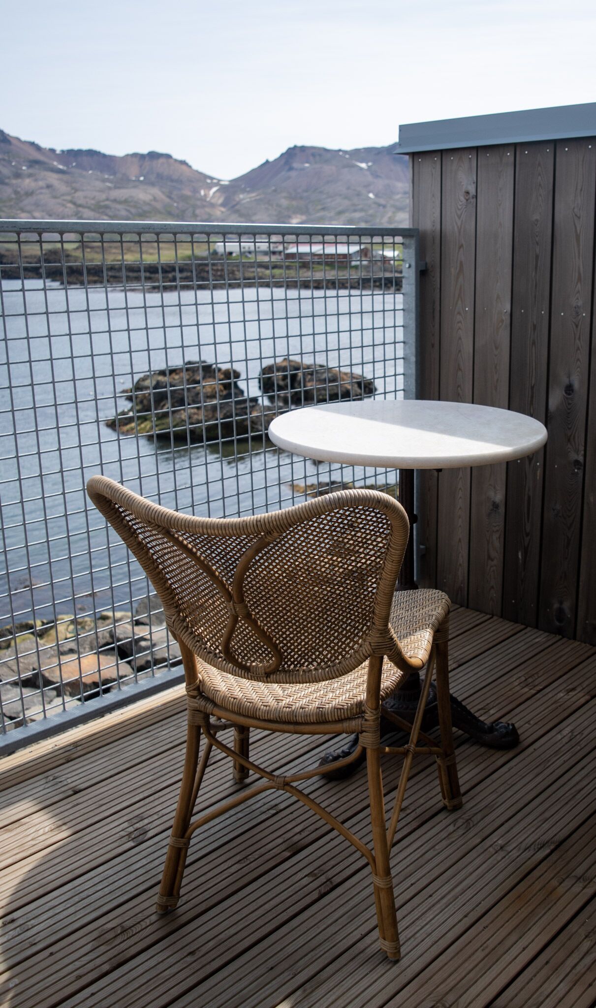 A wicker chair and a round table on a wooden deck overlooking a body of water with mountains in the background.