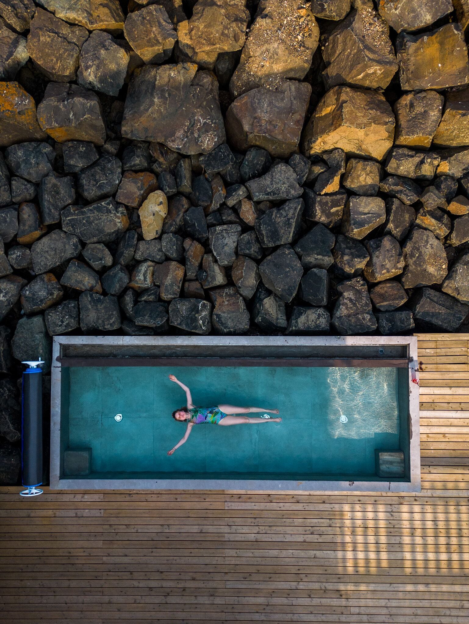 A rectangular swimming pool with a person lying on the edge, surrounded by a wooden deck and a stone wall.