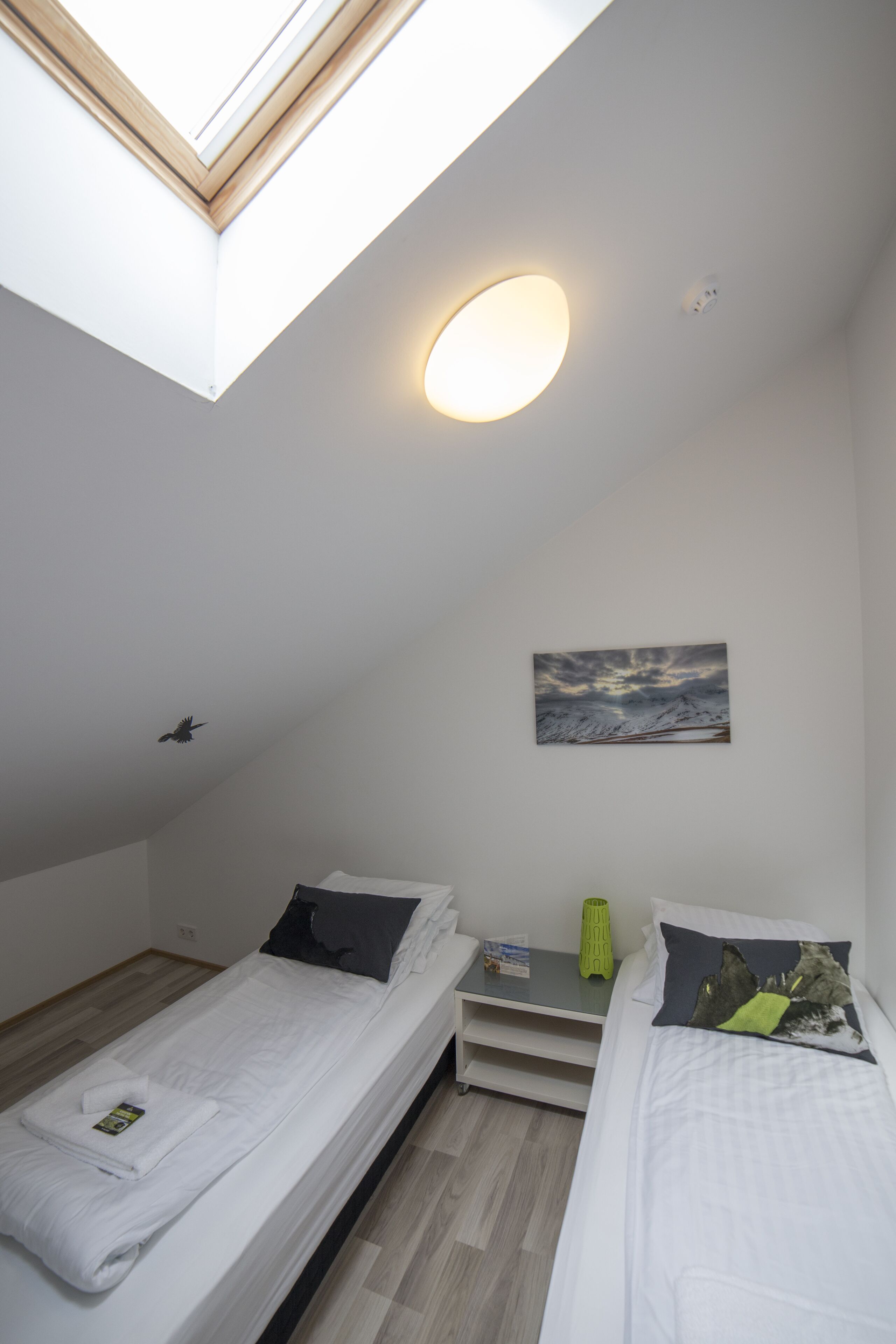 A small, modern room with two single beds, a skylight, a painting, and a bedside table with a lamp.