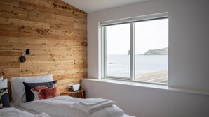 Un dormitorio con pared de madera, una cama con ropa de cama blanca, una mesita de noche con lámpara y vista al mar a través de una ventana.