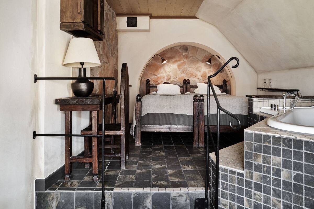 A bedroom with a stone headboard, a bedside table with a lamp, a wooden chair, and a bathroom with a bathtub and tiled walls.