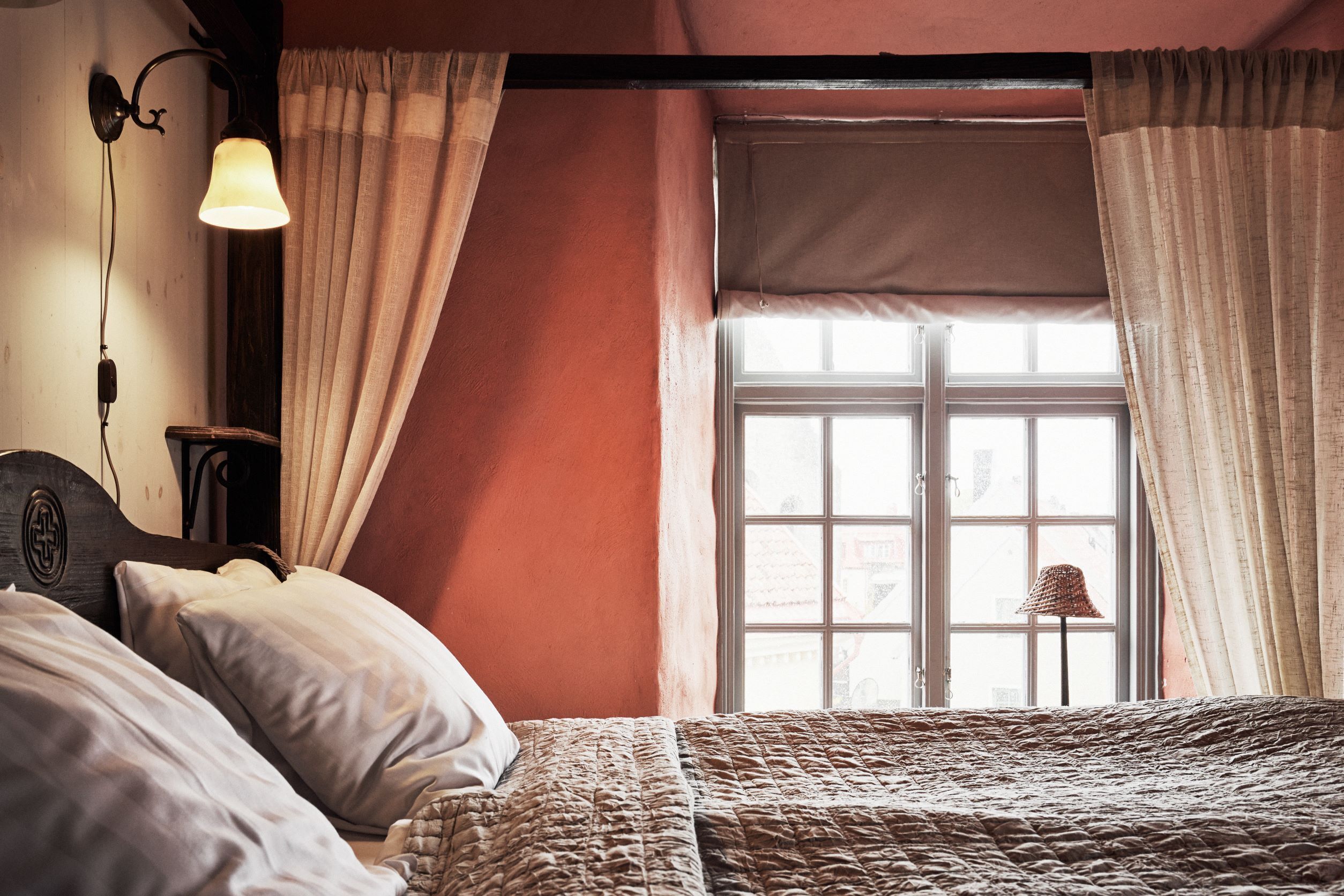 A room with a bed, a window with curtains, a lamp, and a wall-mounted light fixture.