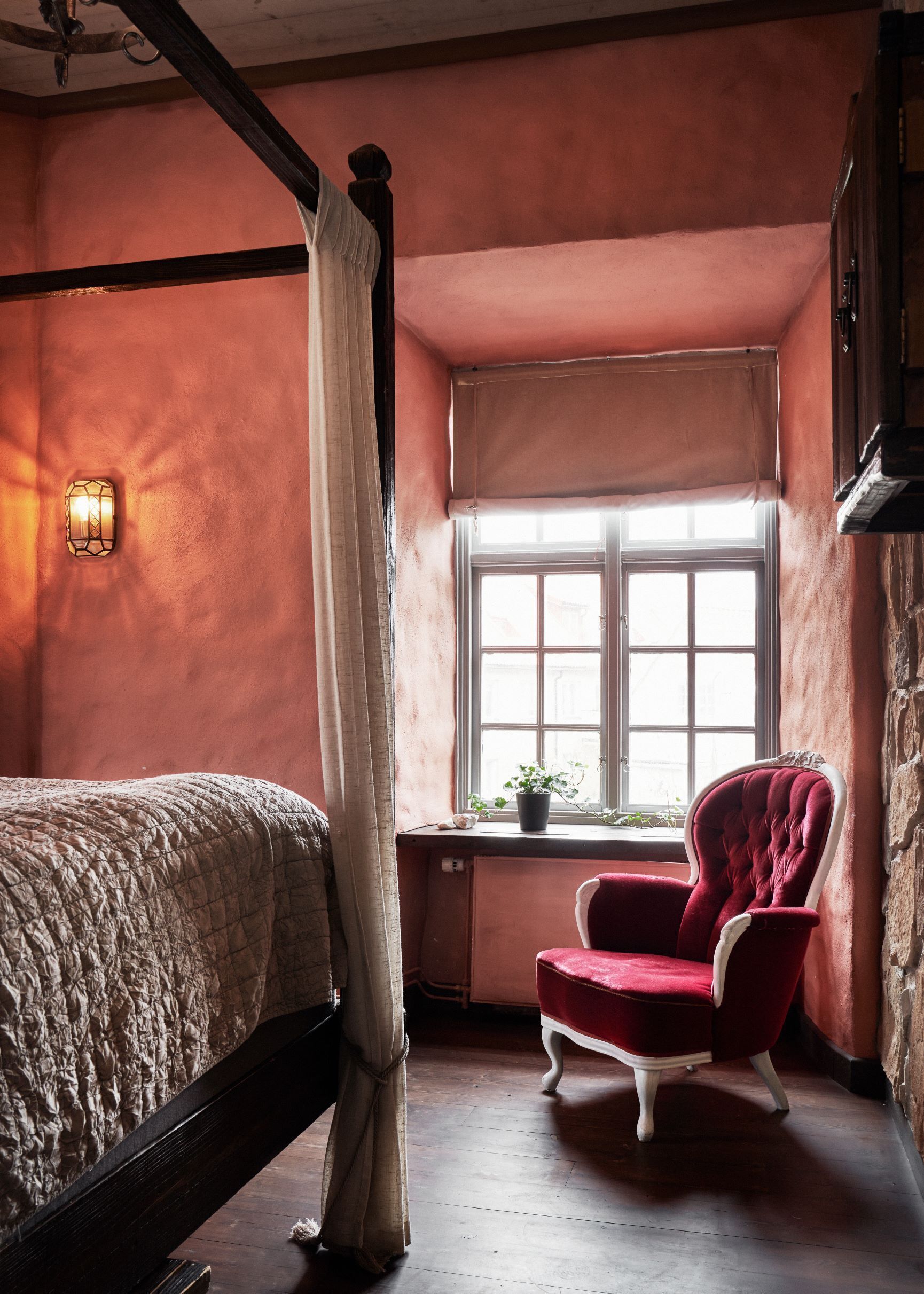 A room with a four-poster bed, a red armchair, a window with a view, and a wall-mounted light fixture.