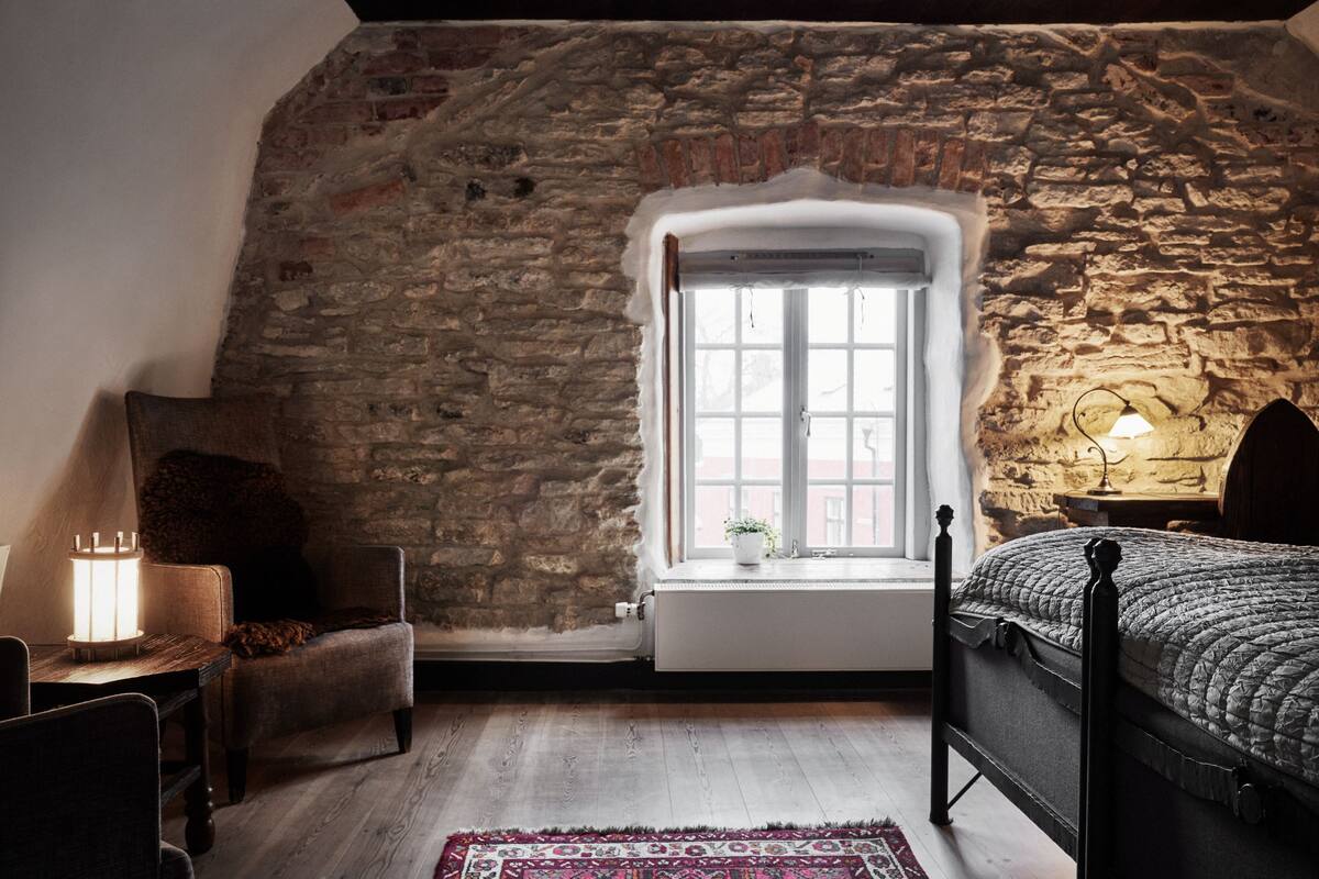 A bedroom with a stone wall, a window, a bed, a chair, a table with a lamp, and a rug.