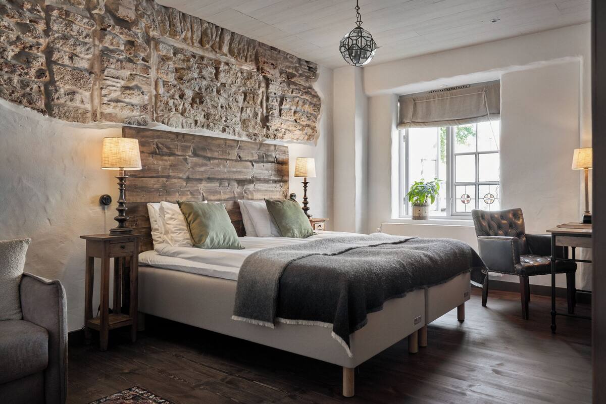 A bedroom with a stone wall, a bed with pillows, a lamp, a chair, and a window with a plant.