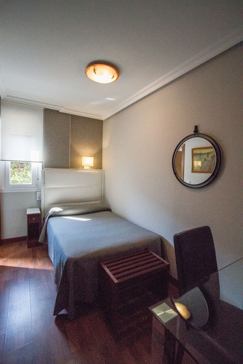 Un dormitorio pequeño y ordenado con cama, mesita de noche, silla y espejo en la pared.