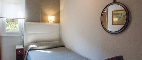 Un dormitorio pequeño y ordenado con cama, mesita de noche, silla y espejo en la pared.
