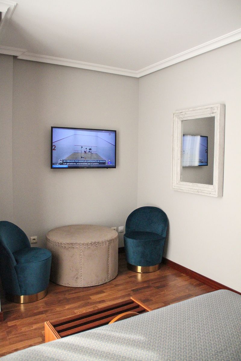 A room with a television mounted on the wall, a round ottoman, two blue chairs, and a bed with a white bedspread.