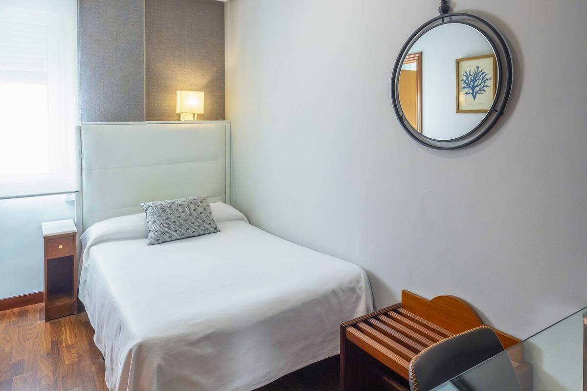 Una habitación de hotel con cama, mesita de noche, silla y un espejo redondo en la pared.