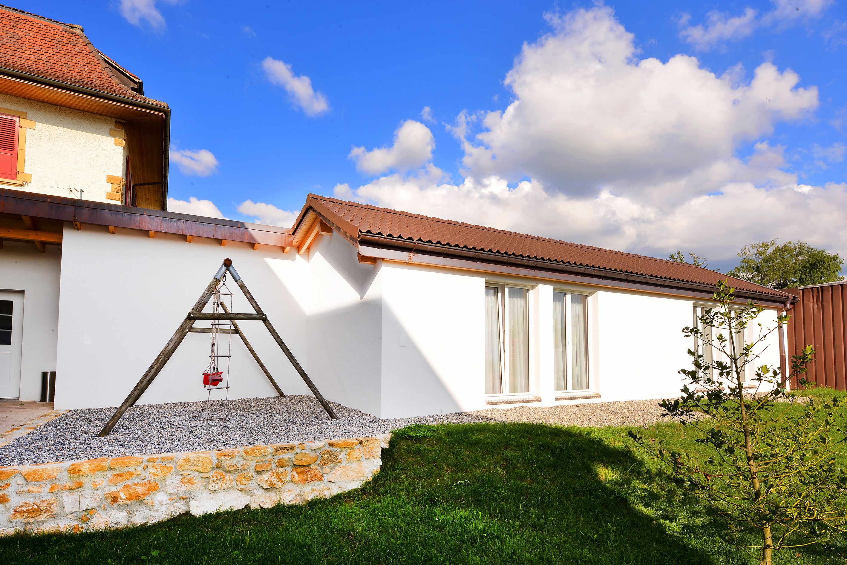 A house with a white exterior, a red roof, and a stone wall in the front yard.