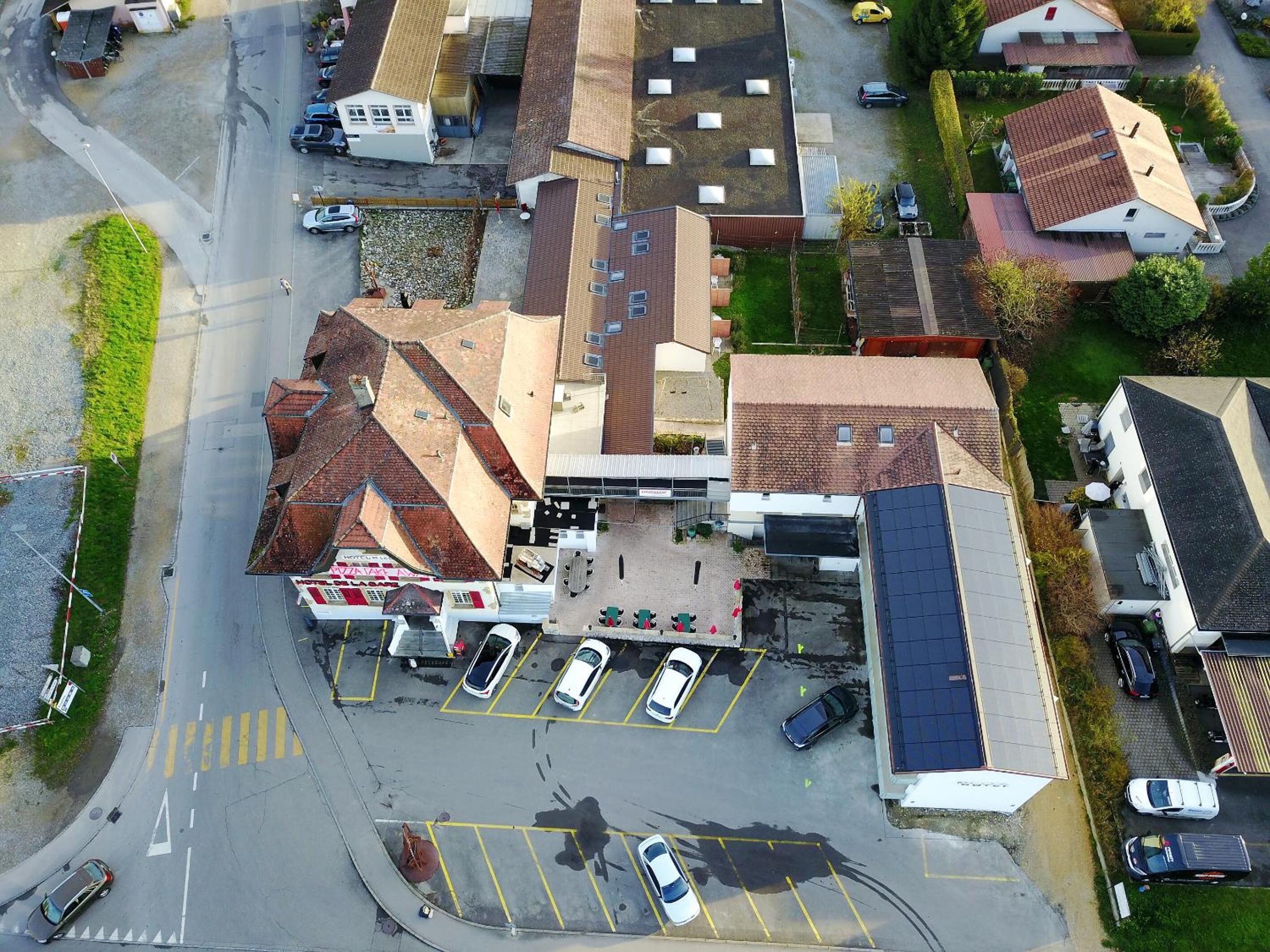 An aerial view of a parking lot with several cars, a building with a sloped roof, and a small green area.