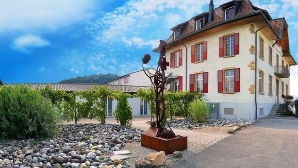 A building with red shutters, a stone facade, and a garden with a metal sculpture.
