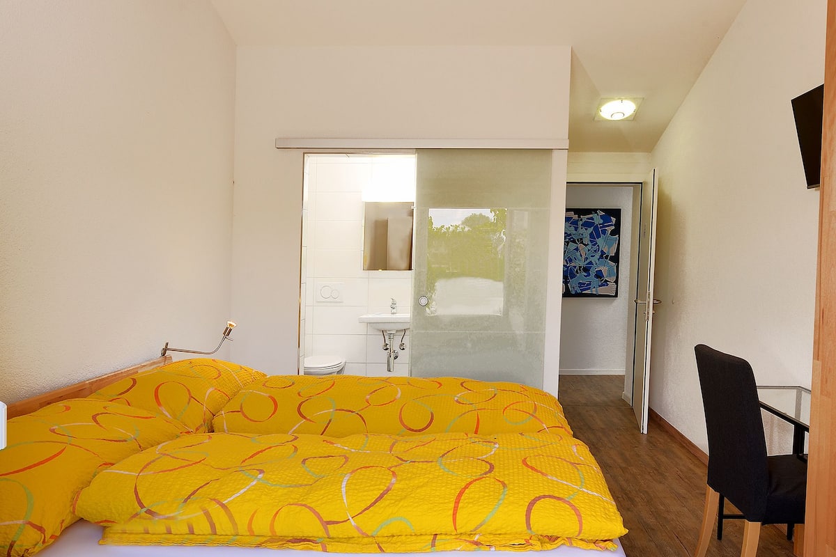 A bedroom with a wooden bed frame, a yellow patterned bedspread, a wooden floor, a black chair, and a white wall with a painting.