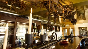 A bar area with a large, ornate metal structure hanging from the ceiling, a bar counter with stools, and a chalkboard menu.