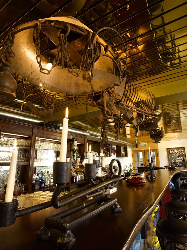A bar area with a large, ornate metal structure hanging from the ceiling, a bar counter with stools, and a chalkboard menu.