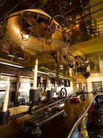 A bar area with a large, ornate metal structure hanging from the ceiling, a bar counter with stools, and a chalkboard menu.