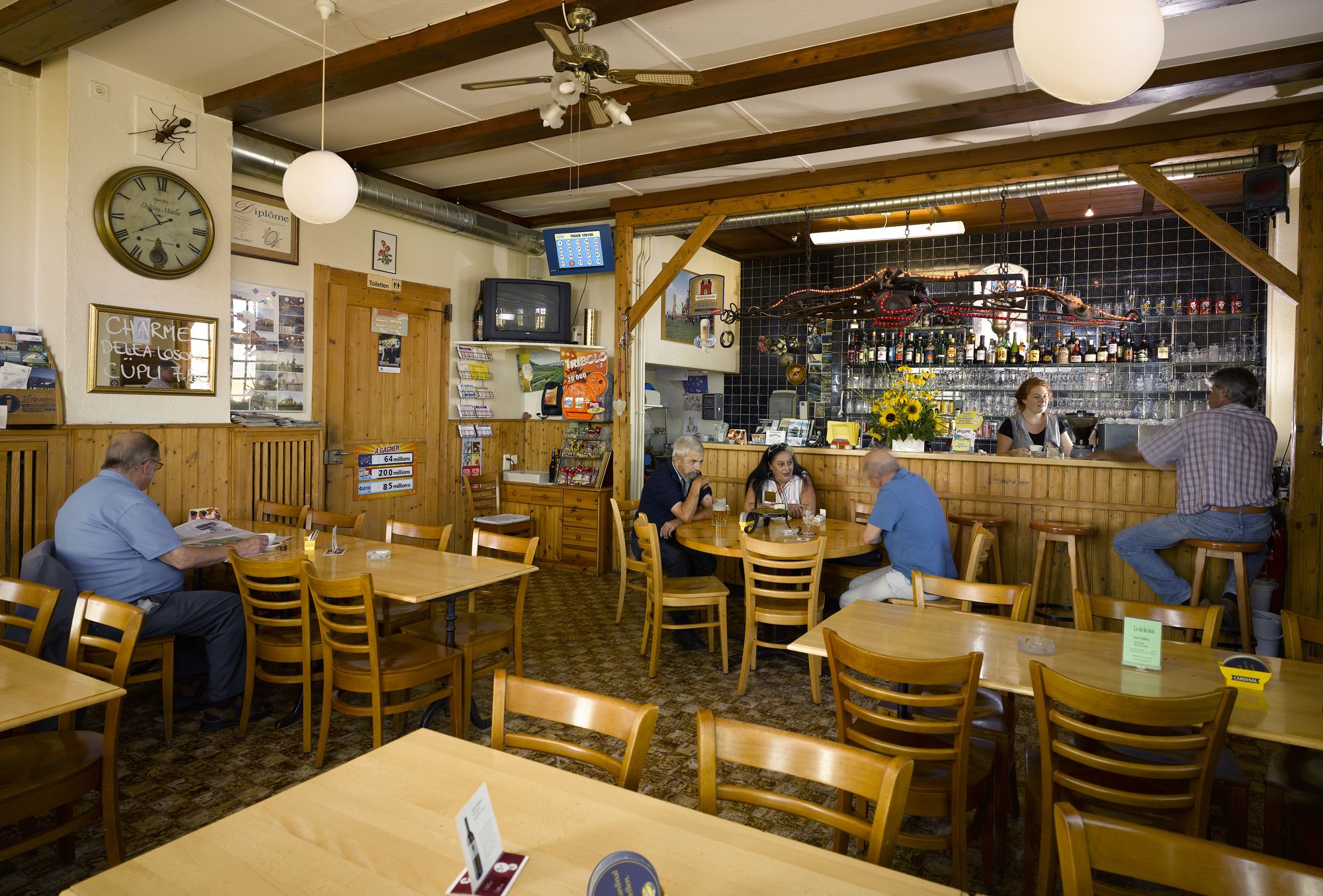 Un restaurante con mesas y sillas de madera, una zona de bar con botellas y un reloj en la pared.
