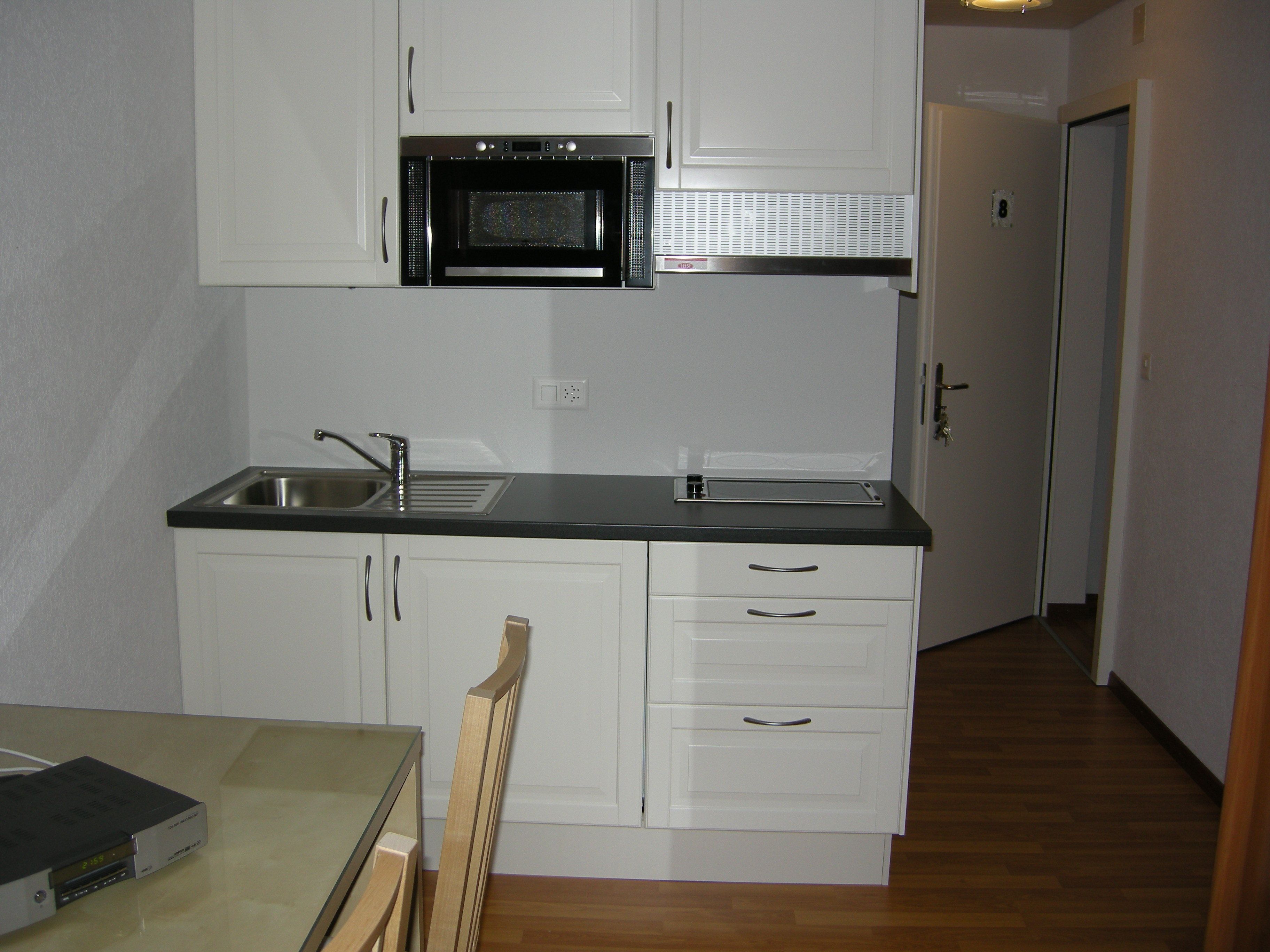 A compact kitchen with white cabinets, a black countertop, a microwave, a sink, and a wooden chair.