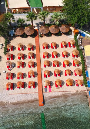 An aerial view of a beachside resort with rows of sun loungers and thatched umbrellas, a wooden walkway, and palm trees. - Butikhan Otel (Bodrum)