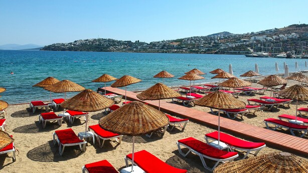 A beach with rows of sun loungers and thatched umbrellas, a clear blue sea, and a hillside town in the background. - Butikhan Otel (Bodrum)