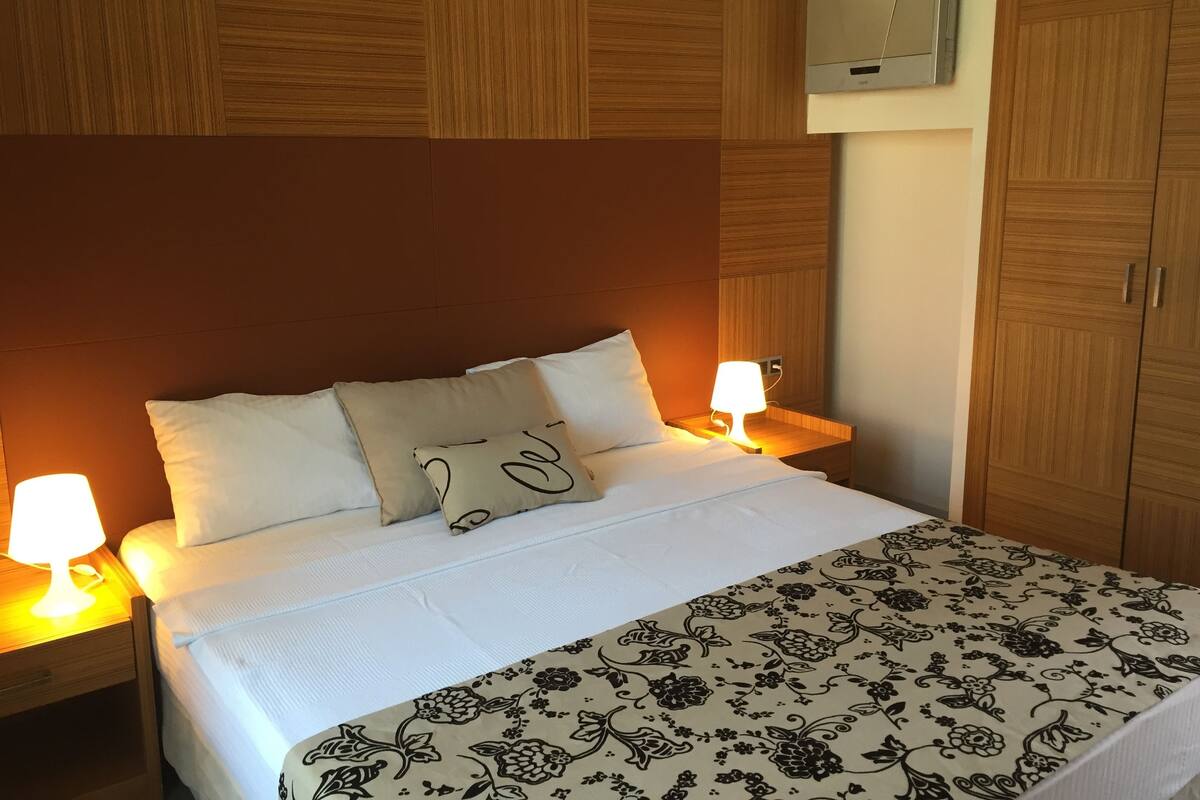 Una cama bien hecha con sábanas blancas y una colcha estampada con motivos florales, flanqueada por dos lámparas de noche sobre mesitas de madera.