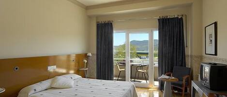 Habitación de hotel con cama, escritorio con televisor y balcón con vistas a las montañas.