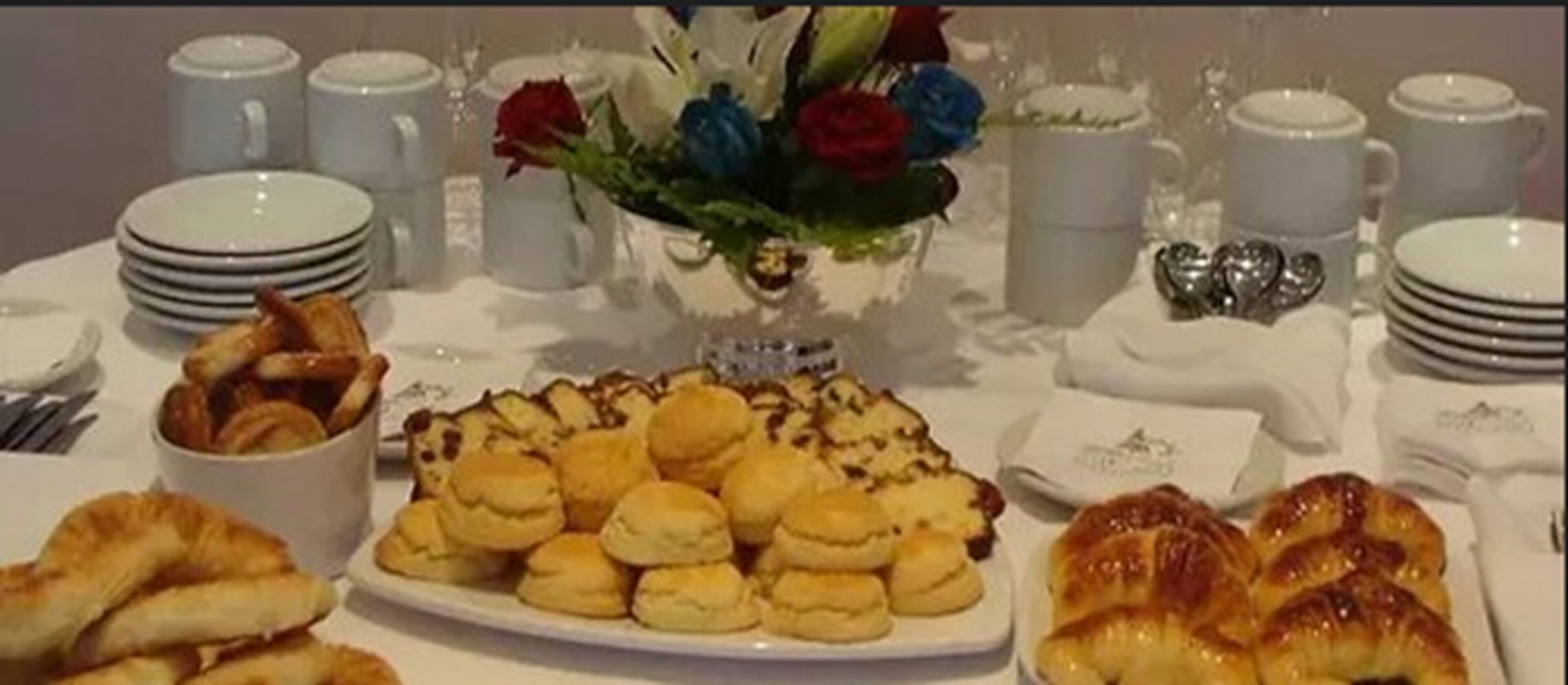 A breakfast buffet with pastries, a vase of flowers, and stacked plates and cups.